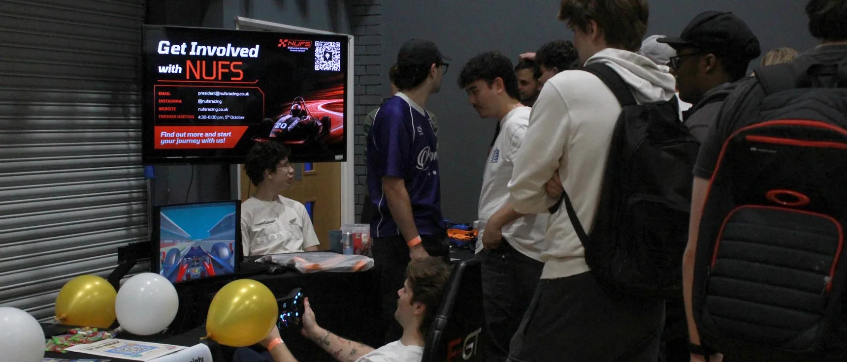 A queue of students at the NUFS stall during the societies fair, with the Get Involved screen and the sim rig
